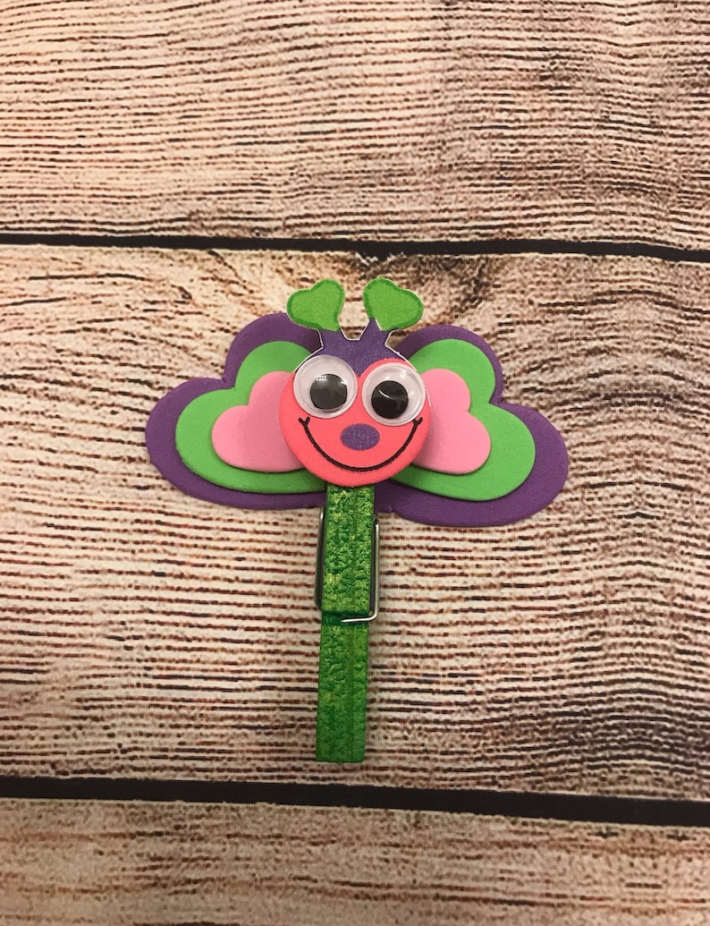 Peut inclure: Une pince &agrave; linge verte avec un motif de papillon rose et vert. Le papillon a des yeux globuleux et une bouche souriante.