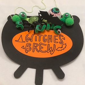 May include: Black cauldron with an orange oval that says "Witches Brew" with a bat graphic. The cauldron is decorated with green pom-poms, googly eyes, a green pipe cleaner, a black spider with glitter, and a black bat with a red eye.