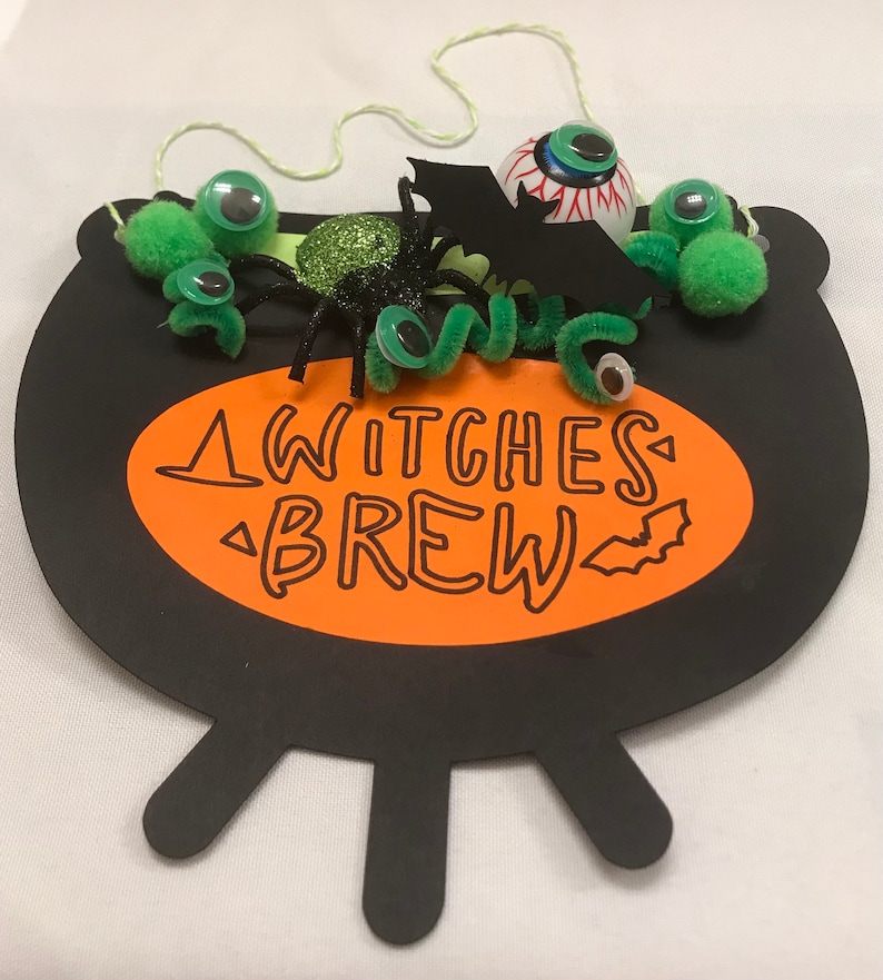 May include: Black cauldron with an orange oval that says "Witches Brew" with a bat graphic. The cauldron is decorated with green pom-poms, googly eyes, a green pipe cleaner, a black spider with glitter, and a black bat with a red eye.