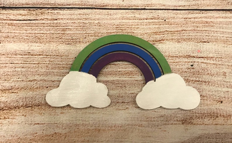 May include: Wooden rainbow with three layers of colour: green, blue, and purple. The rainbow is attached to two white wooden clouds.