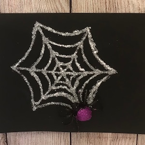 May include: A black cardstock with a silver glitter spider web and a purple glitter spider in the center.