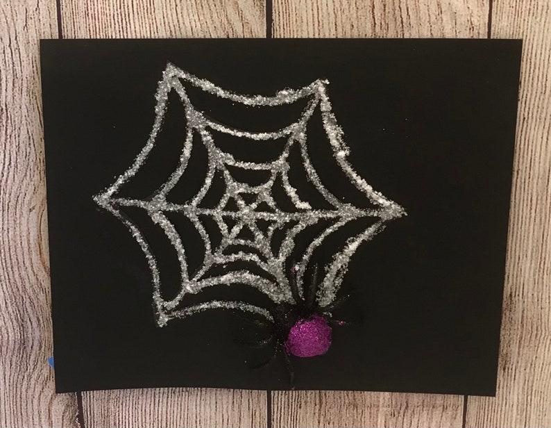 May include: A black cardstock with a silver glitter spider web and a purple glitter spider in the center.