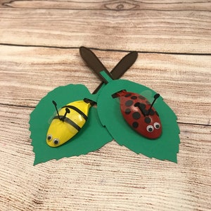 Peut inclure: Deux cuill&egrave;res en plastique peintes pour ressembler &agrave; une coccinelle et une abeille, pos&eacute;es sur des feuilles de papier vert. La coccinelle est rouge avec des points noirs et des yeux globuleux. L'abeille est jaune avec des rayures noires et des yeux globuleux.