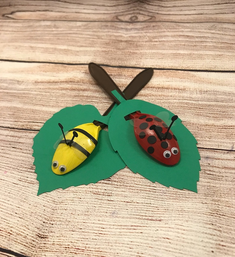 Peut inclure: Deux cuill&egrave;res en plastique peintes pour ressembler &agrave; une coccinelle et une abeille, pos&eacute;es sur des feuilles de papier vert. La coccinelle est rouge avec des points noirs et des yeux globuleux. L'abeille est jaune avec des rayures noires et des yeux globuleux.