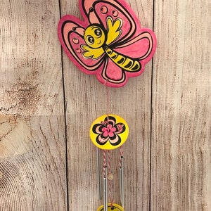 Peut inclure: Un carillon en forme de papillon rose et jaune avec un motif de fleur et de champignon. Le papillon a un visage souriant et des rayures noires et jaunes. Le carillon est suspendu &agrave; une ficelle.