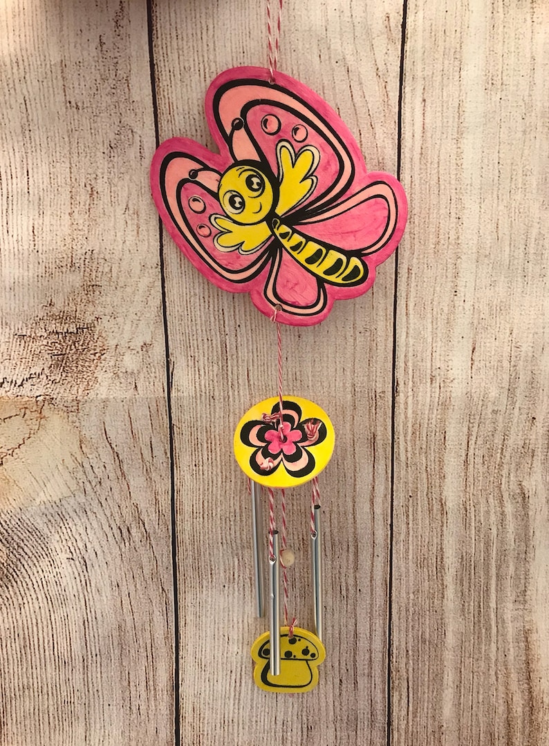 Peut inclure: Un carillon en forme de papillon rose et jaune avec un motif de fleur et de champignon. Le papillon a un visage souriant et des rayures noires et jaunes. Le carillon est suspendu &agrave; une ficelle.