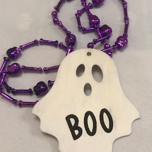 May include: A white ghost-shaped wooden ornament with the word "BOO" written in black. The ornament is attached to a purple beaded necklace with skull-shaped beads.