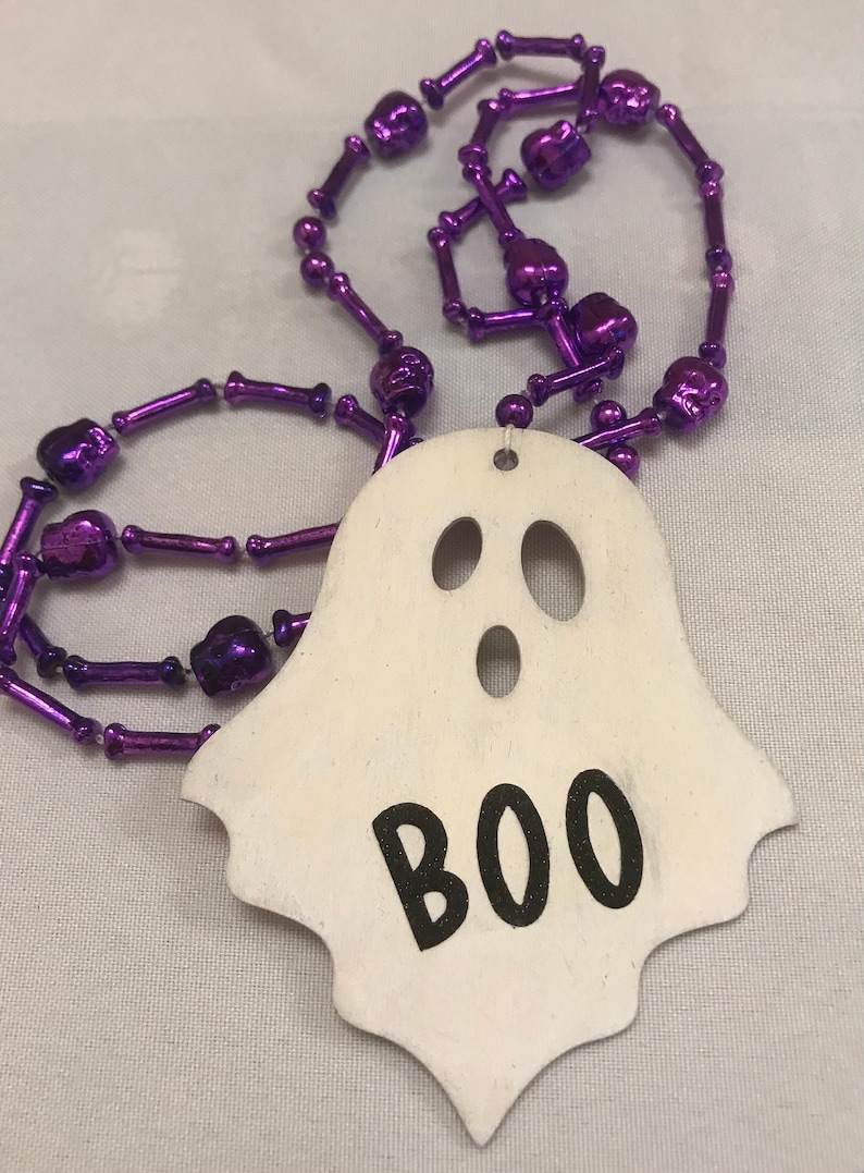 May include: A white ghost-shaped wooden ornament with the word "BOO" written in black. The ornament is attached to a purple beaded necklace with skull-shaped beads.
