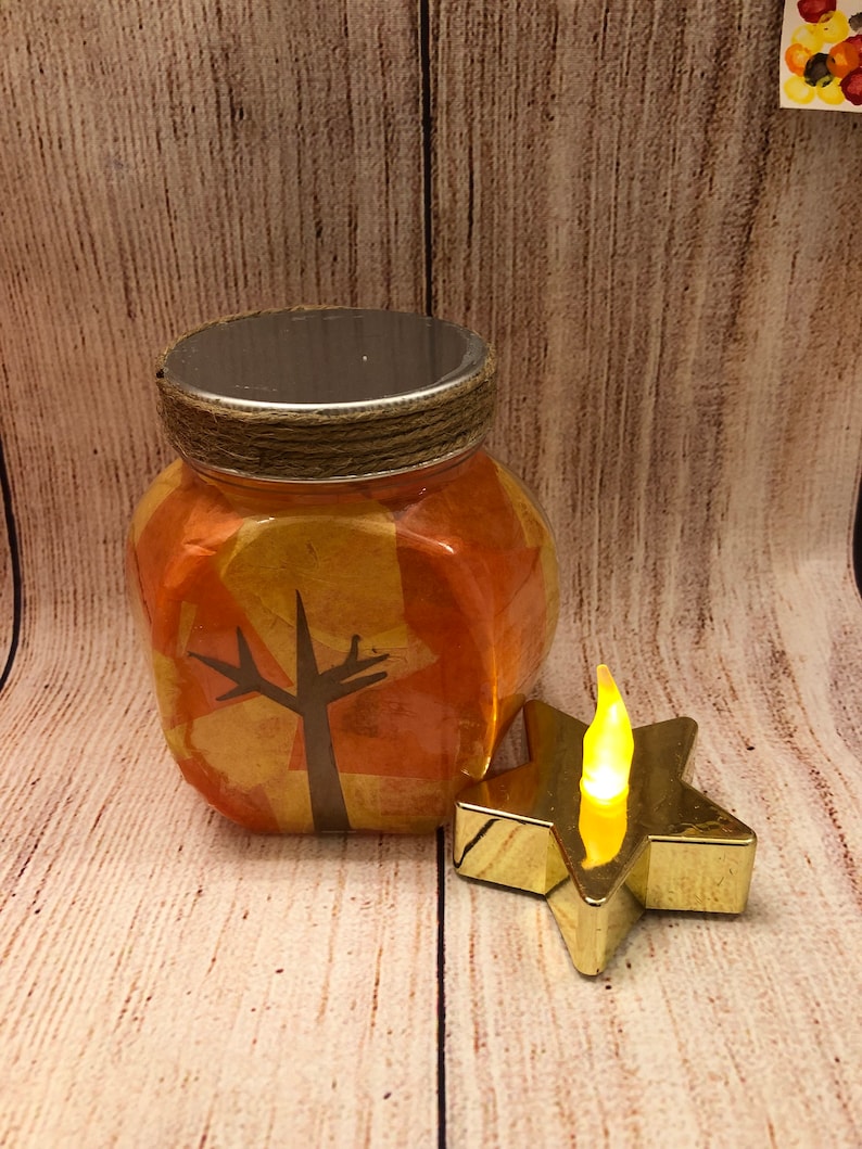 May include: A glass jar with a gold and orange patterned design, featuring a silhouette of a tree. The jar has a silver lid and is tied with twine. A small gold star-shaped LED light sits next to the jar.