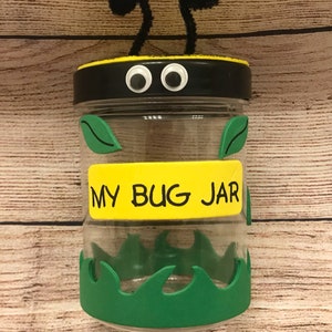 Peut inclure: Un bocal en verre transparent avec un couvercle noir et des yeux globuleux. Le bocal est d&eacute;cor&eacute; de feuilles et d'herbe vertes, et d'une &eacute;tiquette jaune qui dit "My Bug Jar".