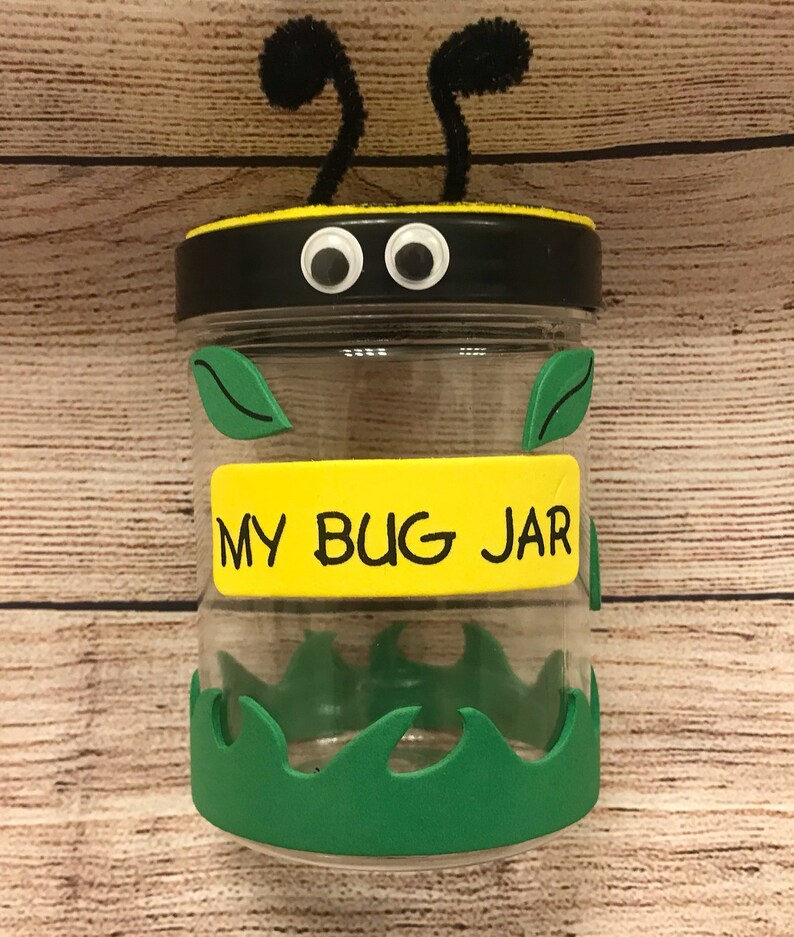Peut inclure: Un bocal en verre transparent avec un couvercle noir et des yeux globuleux. Le bocal est d&eacute;cor&eacute; de feuilles et d'herbe vertes, et d'une &eacute;tiquette jaune qui dit "My Bug Jar".
