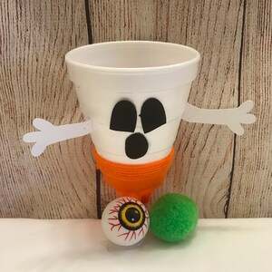 May include: A white styrofoam cup decorated as a ghost with black eyes and a mouth. The ghost has orange yarn wrapped around the bottom and white paper arms. The ghost is sitting on a white surface with a red and yellow eyeball and a green pom-pom.
