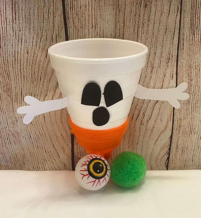 May include: A white styrofoam cup decorated as a ghost with black eyes and a mouth. The ghost has orange yarn wrapped around the bottom and white paper arms. The ghost is sitting on a white surface with a red and yellow eyeball and a green pom-pom.