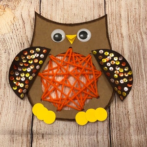 May include: A brown cardboard owl with orange yarn strung through holes in the body. The owl has large googly eyes, a yellow beak, and brown wings decorated with gold and brown sequins.