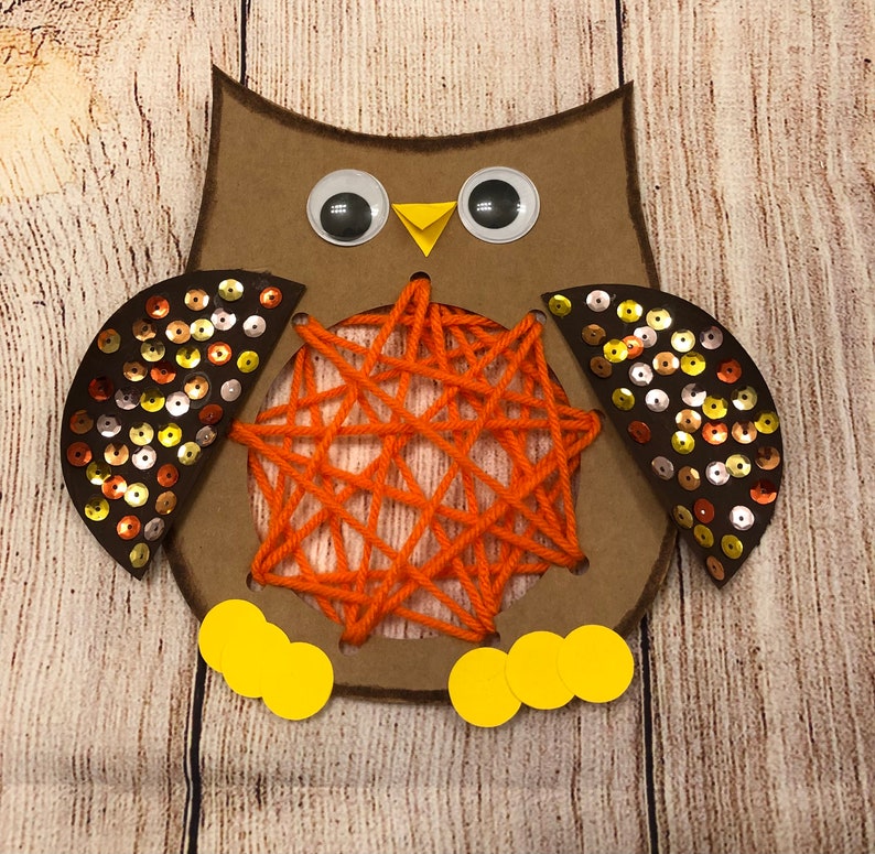 May include: A brown cardboard owl with orange yarn strung through holes in the body. The owl has large googly eyes, a yellow beak, and brown wings decorated with gold and brown sequins.