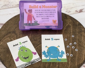 Peut inclure: Un contenant en plastique violet avec une carte à l'intérieur qui dit "Construire un monstre". La carte contient des instructions pour un jeu qui consiste à placer des yeux mobiles sur des cartes de monstres. Il y a deux cartes de monstres, une verte et une bleue, avec le texte "Ajouter 2 yeux" et "Ajouter 5 yeux". Il y a aussi des yeux mobiles éparpillés sur une surface en bois.
