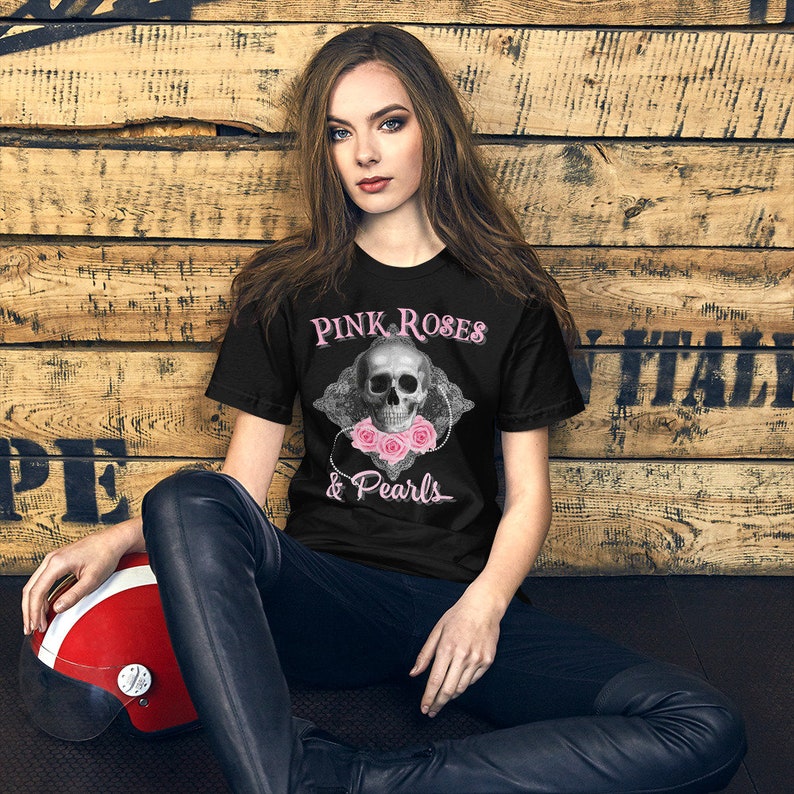 May include: Black t-shirt featuring the text "Pink Roses & Pearls" above a skull graphic adorned with pink roses and a pearl necklace. The shirt is worn by a person sitting on a dark surface.