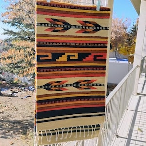 May include: A rectangular woven wall hanging with a cream-colored fringe. The design features horizontal stripes in shades of orange, brown, and black, with repeating arrow-like patterns. The rug is hanging outside on a sunny day.