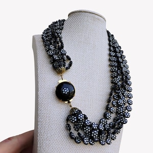 Puede incluir: Un collar vintage de lunares blancos y negros con un gran colgante negro. El collar se exhibe en un maniquí y presenta múltiples hilos de cuentas. El colgante es redondo con un engaste dorado.