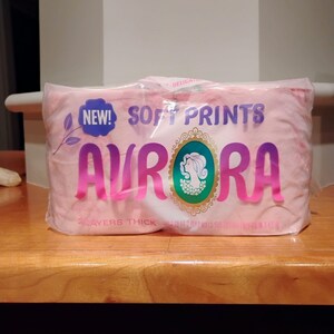 May include: A package of pink Aurora brand soft print tissues. The packaging features the word "AURORA" in large, pink letters, with "NEW!" and "SOFT PRINTS" above. A teal and gold emblem is also visible.