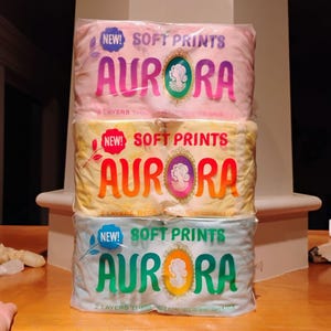 May include: Three packages of Aurora Soft Prints toilet paper in pink, yellow, and blue. Each package has the word "AURORA" in large letters, with the text "SOFT PRINTS" above and "2 LAYERS THICK" below. The packages are stacked on top of each other.