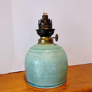 May include: A vintage oil lamp with a round, light-green ceramic base and a dark metal top. The lamp features a brass collar and a wick. The ceramic base has a crackle glaze finish, adding to its antique appearance. The lamp is designed to sit on a flat surface.