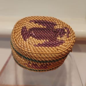 May include: A small, round woven basket with a lid. The basket is made of natural-colored straw with a purple geometric design on the top. A green band encircles the base. The basket is likely handmade.