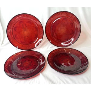 May include: A set of four round, deep red glass plates featuring a pressed geometric diamond pattern along the outer rim and a central starburst design with radiating petal-like motifs. The plates have a smooth, glossy finish.