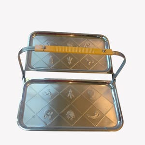 May include: A two-tiered rectangular serving tray made of polished stainless steel with a yellow plastic handle. Each tray features a diamond-patterned surface embossed with various food icons, including a pineapple, fish, and grapes.