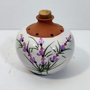 May include: A round ceramic vessel with a white glazed body featuring hand-painted purple lavender sprigs and green stems. The top section is unglazed terracotta with a series of small circular holes and a central opening sealed with a cork stopper.
