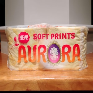 May include: A package of Aurora brand toilet paper. The packaging is yellow with the words "SOFT PRINTS" and "AURORA" in orange and red lettering. A pink oval with a woman's silhouette is also on the packaging. The package is sealed in clear plastic.