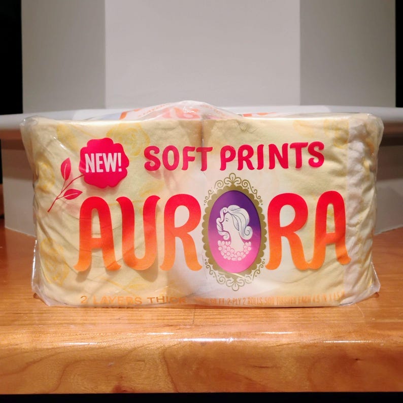 May include: A package of Aurora brand toilet paper. The packaging is yellow with the words "SOFT PRINTS" and "AURORA" in orange and red lettering. A pink oval with a woman's silhouette is also on the packaging. The package is sealed in clear plastic.