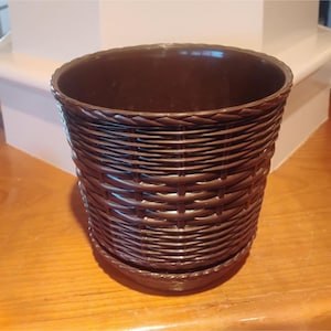 May include: A brown, woven-style plant pot with a matching saucer. The pot has a round shape with a textured, basket-weave design. The rim of the pot has a braided detail. The pot is sitting on a wooden surface.