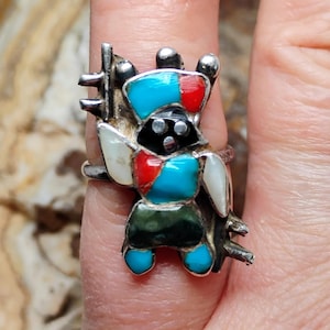 May include: A silver ring featuring a colorful inlaid design. The ring's central figure is a stylized person with a black face, turquoise, red, and white accents. The ring has a unique, artistic appearance.