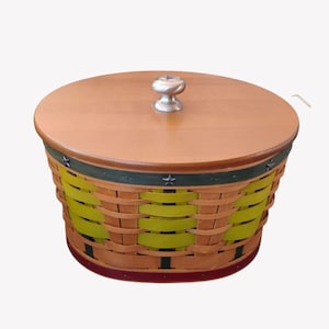 May include: A round woven basket with a wooden lid. The basket has a light brown and yellow weave with a green band and star accents. The lid has a silver knob.
