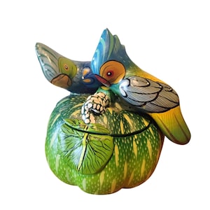 May include: A hand-painted decorative lidded container shaped like a green pumpkin with white speckles and a prominent green leaf. Two colourful birds with blue, yellow, and green plumage are perched on top of the lid, which features a white stem.