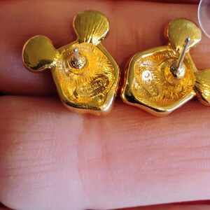 May include: A pair of gold-colored stud earrings in a Mickey Mouse design. The earrings have a post backing and a textured surface. The back of the earrings are visible, with the word "Disney" stamped on the surface.