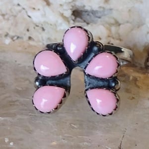 Könnte beinhalten: Ein Silberring mit fünf tropfenförmigen, hellrosa Steinen. Jeder Stein ist in einer gezackten Fassung gefasst. Der Ring hat ein offenes Design, wobei die Steine in einem blütenartigen Muster angeordnet sind. Der Ring ist vor einem neutralen Hintergrund platziert.