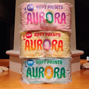May include: Three packages of "AURORA" brand soft prints, each wrapped in clear plastic. The top package is pink, the middle is yellow, and the bottom is light blue. Each package has the words "NEW! SOFT PRINTS" and "AURORA" printed on it.
