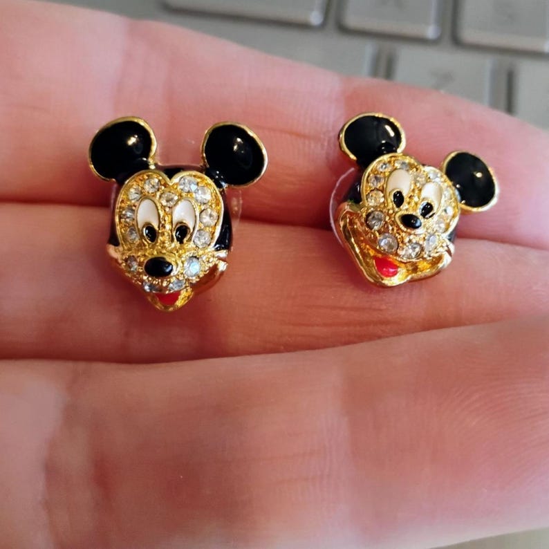 May include: A pair of gold-toned Mickey Mouse stud earrings. Each earring features a black enamel head with sparkling crystal accents, white eyes, a black nose, and a red mouth. The earrings are held in a person's hand.