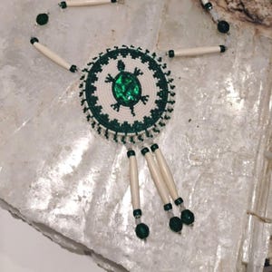 May include: A beaded necklace featuring a central circular pendant with a turtle design. The pendant is white and green, with a large green gem in the center. The necklace has off-white tubular beads and green accents. The necklace is on a white surface.