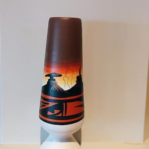 May include: A decorative wooden bottle stopper with a brown top and a white base. The stopper features a painted landscape scene with a sunset, mountains, and geometric designs in black and orange.