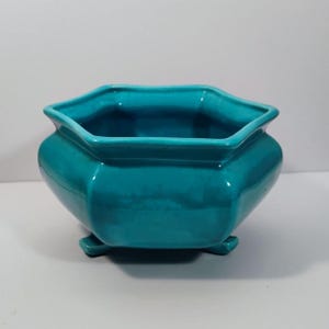 May include: A glossy, turquoise ceramic planter with a hexagonal shape and a wide, open top. The vessel features a smooth, reflective finish and is supported by four small, integrated feet at the base, creating a stable and elevated appearance.