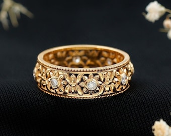 May include: A gold band ring with a delicate floral design and small clear gemstones. The ring is on a black fabric background.