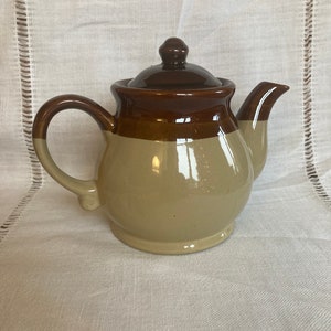 May include: A brown and beige ceramic teapot with a lid. The teapot has a rounded body and a spout on the side.