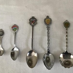 May include: Five silver souvenir spoons with different designs. Each spoon has a different design on the handle, including a castle, a mountain, a flower, and a coat of arms. The spoons are all in good condition and would make a great addition to any collection.