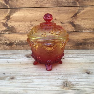 May include: A vintage glass candy dish with a red and yellow ombre finish. The dish is decorated with a grapevine pattern and has three legs.