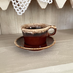 May include: A brown ceramic coffee cup and saucer set. The cup has a rounded shape with a handle and a dark brown rim. The saucer is a matching brown color. The set is displayed on a light-colored surface.