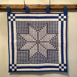 May include: A blue and white quilted wall hanging with a star design. The star is centered on a blue and white gingham background. The border is blue and white with a checkered pattern in the corners. The wall hanging is suspended from a wooden rod.