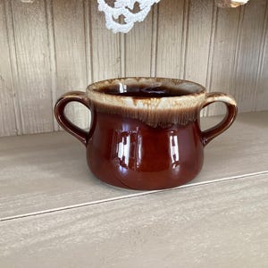 May include: A glossy, brown ceramic mug with two handles. The rim has a light brown, dripped glaze. The mug sits on a light-colored surface. The mug is likely for serving soup or beverages.